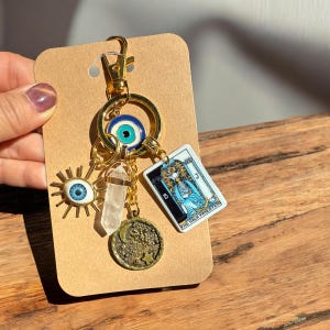 Peut inclure: Porte-cl&eacute;s en or avec une vari&eacute;t&eacute; de breloques, dont un cristal transparent, une lune croissante, une &eacute;toile, un mauvais &oelig;il bleu et une carte de tarot avec le texte "La Grande Pr&ecirc;tresse".