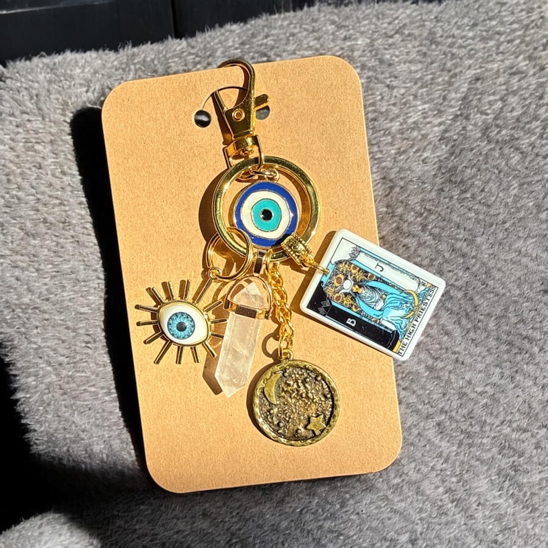 Peut inclure: Porte-cl&eacute;s en or avec une vari&eacute;t&eacute; de breloques, dont un &oelig;il bleu mal&eacute;fique, un cristal transparent, un soleil avec un &oelig;il bleu, une lune avec des &eacute;toiles et une carte de tarot avec le texte "The Star".