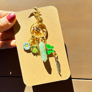 Könnte beinhalten: Ein goldfarbener Schlüsselanhänger mit mehreren Anhängern. Zu den Anhängern gehören eine Biene, ein grüner Kristall, ein grüner Schmetterling, eine blau-grüne Blume und eine Feder. Der Schlüsselanhänger ist an einer braunen Karte befestigt.