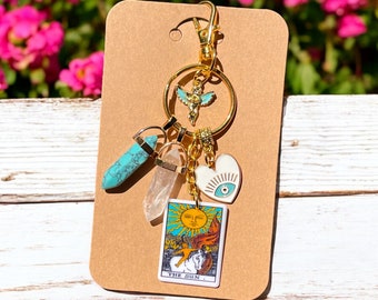 May include: Gold keychain with a turquoise stone, a clear crystal, a white heart with a blue eye, and a tarot card charm that says "The Sun".