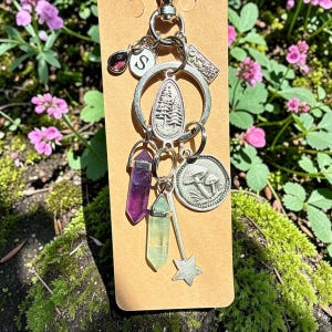Peut inclure: Un porte-clés argenté composé d'un anneau circulaire, d'une breloque en forme de goutte avec une gravure de sapin, d'un disque avec un motif de champignon, deux pointes cristallines violette et vert clair, une étoile et une lettre S.