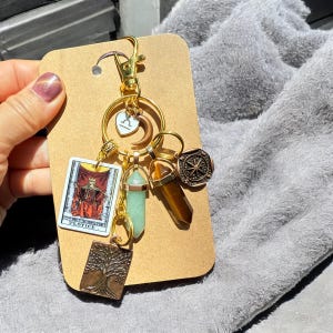May include: Gold keychain with a variety of charms, including a tarot card, a compass, a crystal, and a tree of life. The tarot card is the Justice card.