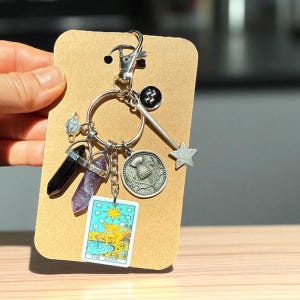 May include: A silver keychain with various charms, including a black crystal, a purple crystal, a coin, a star, and a tarot card. The tarot card features an illustration of a person in water with the text "THE STAR". The keychain is attached to a brown cardboard backing.