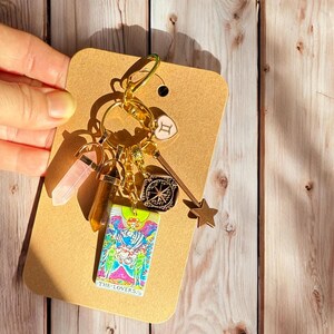May include: Gold keychain with a variety of charms, including a tarot card with the image "The Lovers", a pink crystal, a brown crystal, a gold compass, a gold star, and a gold heart with the Gemini zodiac symbol.