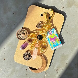 May include: A gold-colored keychain with various charms, including a crescent moon, a coin with a bird design, a compass, and two crystal pendants. A tarot card with a colorful illustration is also attached. The keychain is on a brown card.