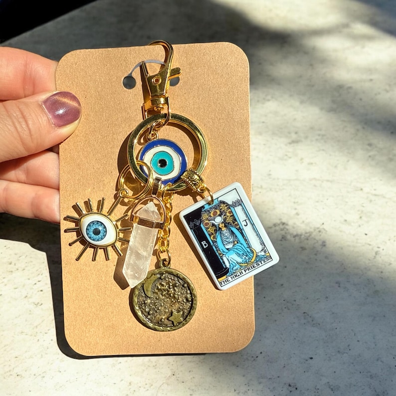 Peut inclure: Porte-cl&eacute;s en or avec un charme &oelig;il bleu mal&eacute;fique, un charme en cristal transparent, un charme de lune dor&eacute;e et un charme de carte de tarot qui dit "La Grande Pr&ecirc;tresse".