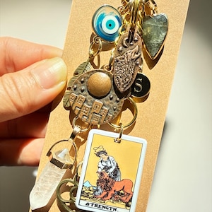 May include: A gold-toned keychain with multiple charms, including a clear quartz crystal, a tarot card with the word "STRENGTH," and a blue and white evil eye. Other charms include a heart, a feather, and a circular charm with a design.