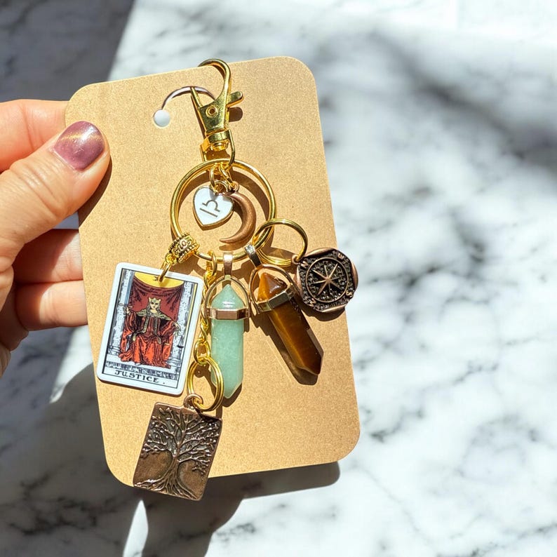 May include: Gold keychain with a variety of charms, including a tarot card, a compass, a crystal, and a tree of life. The tarot card is the Justice card.