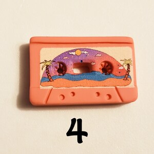 May include: Pink plastic cassette tape charm featuring a printed image of a beach scene with palm trees, a sunset, and ocean waves.