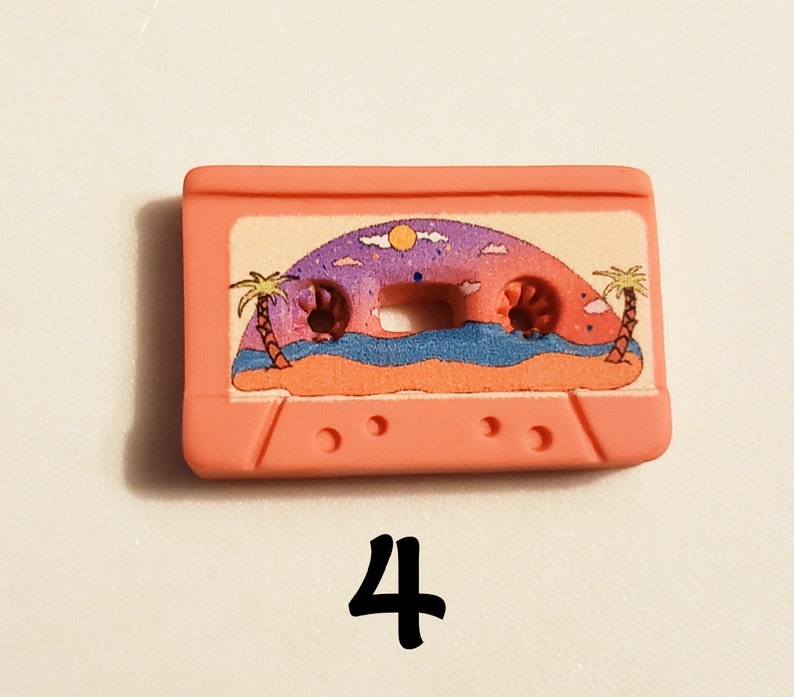 May include: Pink plastic cassette tape charm featuring a printed image of a beach scene with palm trees, a sunset, and ocean waves.