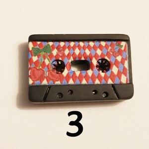 May include: Black cassette tape button with a red, white and blue chequered pattern and a cherry design. The button has two holes on the bottom.