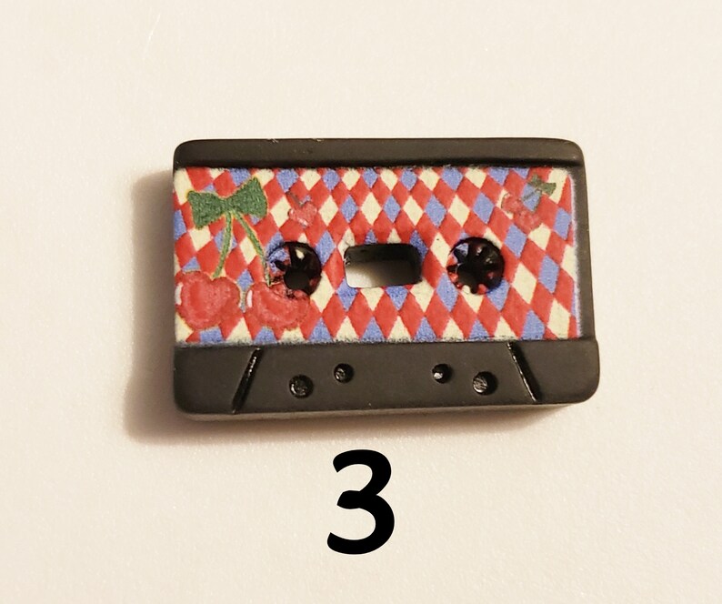 May include: Black cassette tape button with a red, white and blue chequered pattern and a cherry design. The button has two holes on the bottom.