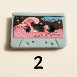 May include: A light blue cassette tape with a pink wave and stars on a black background. The tape has a white cloud in the top right corner.
