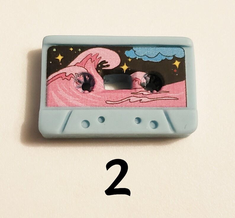 May include: A light blue cassette tape with a pink wave and stars on a black background. The tape has a white cloud in the top right corner.