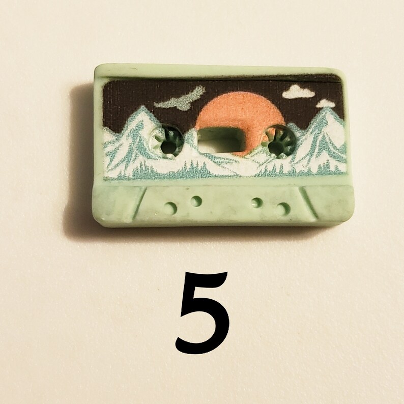 May include: A light green cassette tape featuring a mountain and sunset design. The tape has a black background with a white mountain range and a pink sunset. The tape has a white border and a black label with the words "Sunset Mountain" printed on it.