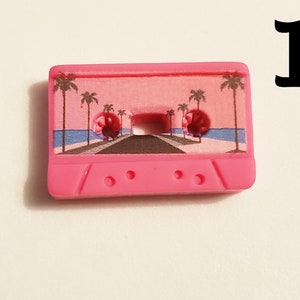 May include: Pink cassette tape charm with a palm tree and beach scene printed on the front. The charm has three small holes on the bottom.