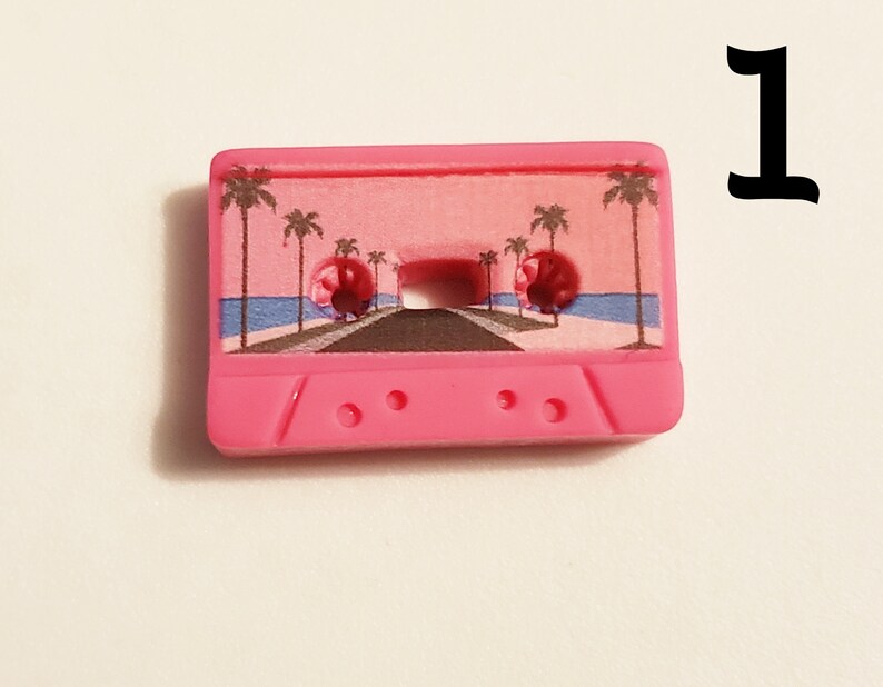 May include: Pink cassette tape charm with a palm tree and beach scene printed on the front. The charm has three small holes on the bottom.