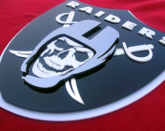 May include: A black and white 3D Raiders logo featuring a skull wearing a helmet and crossed swords. The logo is on a black background with a white border.