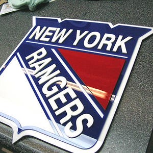 Puede incluir: Un logotipo blanco y azul de los New York Rangers con un triángulo rojo en el centro. El logotipo está hecho de plástico y tiene un efecto 3D.