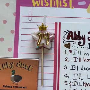 Può includere: Una candela a forma di stella con il numero 24 e una corona d'oro, su un bastoncino di legno. La candela è su uno sfondo rosa con le parole "Magical Wishlist". È visibile anche un'insegna di un ristorante cinese.
