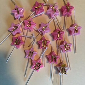 K&ouml;nnte beinhalten: Rosa sternf&ouml;rmige Kerzen mit goldenen Zahlen von 1 bis 16. Die Kerzen sind auf Holzstielen und in einem zuf&auml;lligen Muster angeordnet.