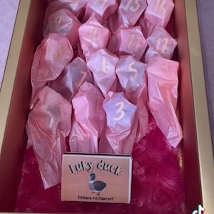K&ouml;nnte beinhalten: Eine rosa Box gef&uuml;llt mit kleinen rosa sternf&ouml;rmigen S&uuml;&szlig;igkeiten, jede mit einer darauf gedruckten Nummer. Die Box ist mit rosa Seidenpapier ausgekleidet. Ein kleines Schild am Boden der Box lautet "Lucky Duck Chinesisches Restaurant."
