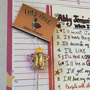 Może przedstawiać: Różowa i biała "Magiczna lista życzeń" z pudełkiem restauracji chińskiej "Lucky Duck" i świecą urodzinową w kształcie gwiazdy "16". Lista zawiera odręczne życzenia.