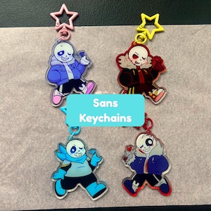 May include: Four acrylic keychains featuring cartoon skeleton characters, each with a star-shaped clasp. The characters are depicted in various colour schemes including blue, red, and light blue, with expressive faces. A light blue rectangle with the text Sans Keychains is centred over the items.