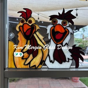 Puede incluir: Dos pollos de vidrio coloreado con ojos grandes y picos abiertos. El pollo de la izquierda es amarillo y marrón, mientras que el pollo de la derecha es blanco con una cresta y una barbilla rojas. El texto "Kim Mergen Glass Design" es visible en la parte inferior de la imagen.