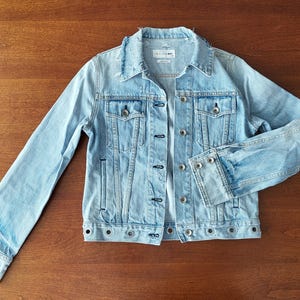 Pode incluir: Jaqueta jeans azul clara com um design clássico. Possui fechos de botão, bolsos no peito e detalhes desgastados na gola e punhos. A jaqueta está colocada sobre uma superfície de madeira.