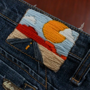 May include: A blue denim jacket with a colorful embroidered patch depicting a sunset scene with a mountain, clouds, and a sun. The patch is sewn onto the back of the jacket.