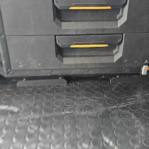 May include: Black storage drawers with yellow accents and handles. A yellow work light is visible on the right. The drawers are part of a larger storage system, likely for tools or equipment. The floor has a textured, rubberised surface.