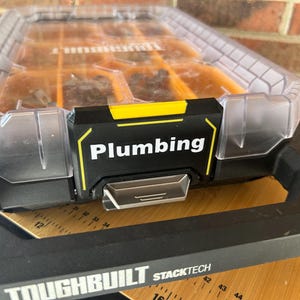 May include: A ToughBuilt StackTech organiser box with a clear lid and orange interior compartments. The black front latch features the white text Plumbing. A metal ruler is visible underneath, showing measurements in centimetres.