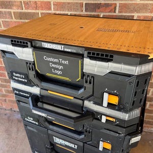 May include: A stack of black and grey Toughbuilt StackTech toolboxes with a wooden top. The boxes have orange latches and handles. Text on the boxes includes "Custom Text Design Logo" and "Batty's Hardware".