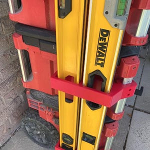 May include: Two yellow DeWalt spirit levels are held vertically by a custom red plastic mounting bracket attached to a red rolling tool box. The levels feature integrated green liquid vials for precision levelling and are secured in place.