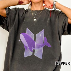 Pode incluir: T-shirt cinzento escuro com um design de baleia roxa dentro de um logótipo estilizado. O design inclui a palavra "PEPPER" em branco no canto inferior direito. A t-shirt tem mangas curtas e gola redonda.