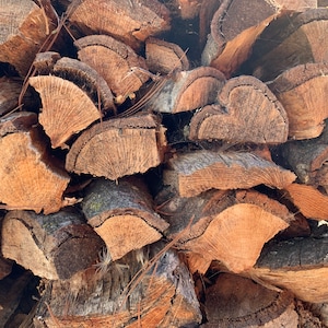 May include: A stack of split firewood logs featuring rough, dark brown bark on the outer edges and light tan, textured wood grain on the interior faces. The logs are cut into various semi-circular and triangular shapes, piled together in a dense arrangement.