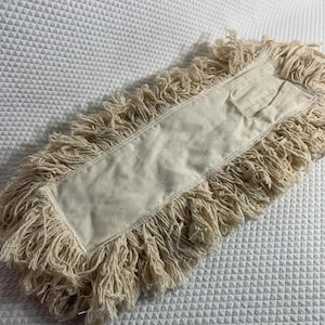 May include: A rectangular dust mop head made of off-white cotton yarn with a thick, fringed border and a central flat fabric panel. The cleaning tool features a reinforced stitched edge and a small pocket-like attachment on one end for mounting.