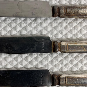 May include: Three table knives with silver-toned stainless steel blades and rectangular handles. Each blade is stamped with the word STAINLESS. The handles feature a weathered, metallic finish with a central rectangular recessed detail.