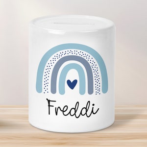 Puede incluir: Hucha de cerámica blanca con un diseño de arcoíris azul y gris y un pequeño corazón azul en el centro. El nombre "Freddi" está escrito en cursiva negra debajo del arcoíris.