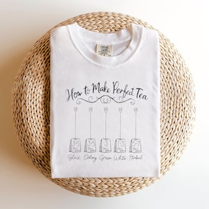 Può includere: T-shirt bianca con la scritta "How to Make Perfect Tea" e un diagramma che mostra i tempi di infusione del tè. Il diagramma include i tempi per tè nero, oolong, verde, bianco ed erbe. La maglietta è piegata su una superficie tessuta.