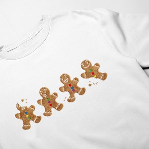 Może przedstawiać: Biała bluza z nadrukiem czterech ludzików z piernika. Piernikowe ludziki mają brązowe ciała, kolorowe guziki z cukierków i uśmiechnięte twarze. Jeden z ludzików z piernika jest częściowo zjedzony, z okruchami.