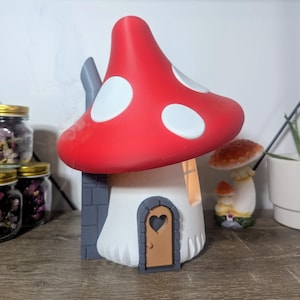 May include: A red and white mushroom-shaped house with a grey brick base and a brown door with a heart-shaped window. The house has a white interior with a light shining through the window. There is smoke coming from the chimney.