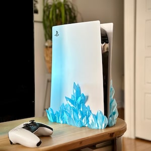 Może przedstawiać: Biała konsola PlayStation 5 z niebieskim stojakiem przypominającym kryształ. Konsola ma logo PlayStation z przodu. Stojak jest wykonany z plastiku drukowanego w 3D i ma imitować kryształy lodu.