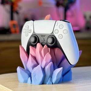 Pode incluir: Um controle branco do PlayStation 5 está em um suporte em forma de cristal azul e rosa. O controle tem botões pretos e um logotipo preto do PlayStation.