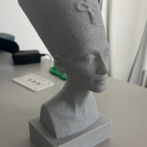 Può includere: Busto grigio di Nefertiti, con una dettagliata raffigurazione dell'antica regina egizia. La scultura ha una superficie strutturata ed è montata su una base rettangolare. Il busto è una rappresentazione dell'arte storica.
