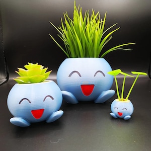 May include: Three blue ceramic planters shaped like cartoon faces with smiling expressions. The planters are filled with green plants.