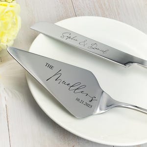 以下が含まれることがあります： 名前と日付が刻まれたシルバーのケーキナイフとサーバーセット。「Stephanie & Daniel」と書かれたナイフと、「The Muellers 10.21.2025」と書かれたサーバー。白いお皿の上にセットされ、背景には黄色の花があります。