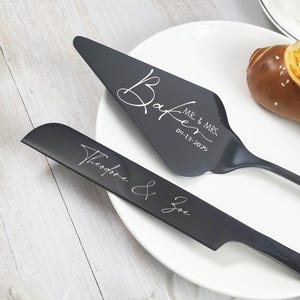 May include: A black cake server and knife set with white script. The server reads "Baker MR. & MRS. 09-13-2025". The knife reads "Theodore & Zoe". The set is on a white plate.