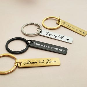Puede incluir: Una colección de llaveros personalizados en oro, plata y negro. Cada llavero presenta texto grabado, incluyendo frases como "Having you is my luck" y nombres con fechas. Los llaveros tienen formas redondas o rectangulares.